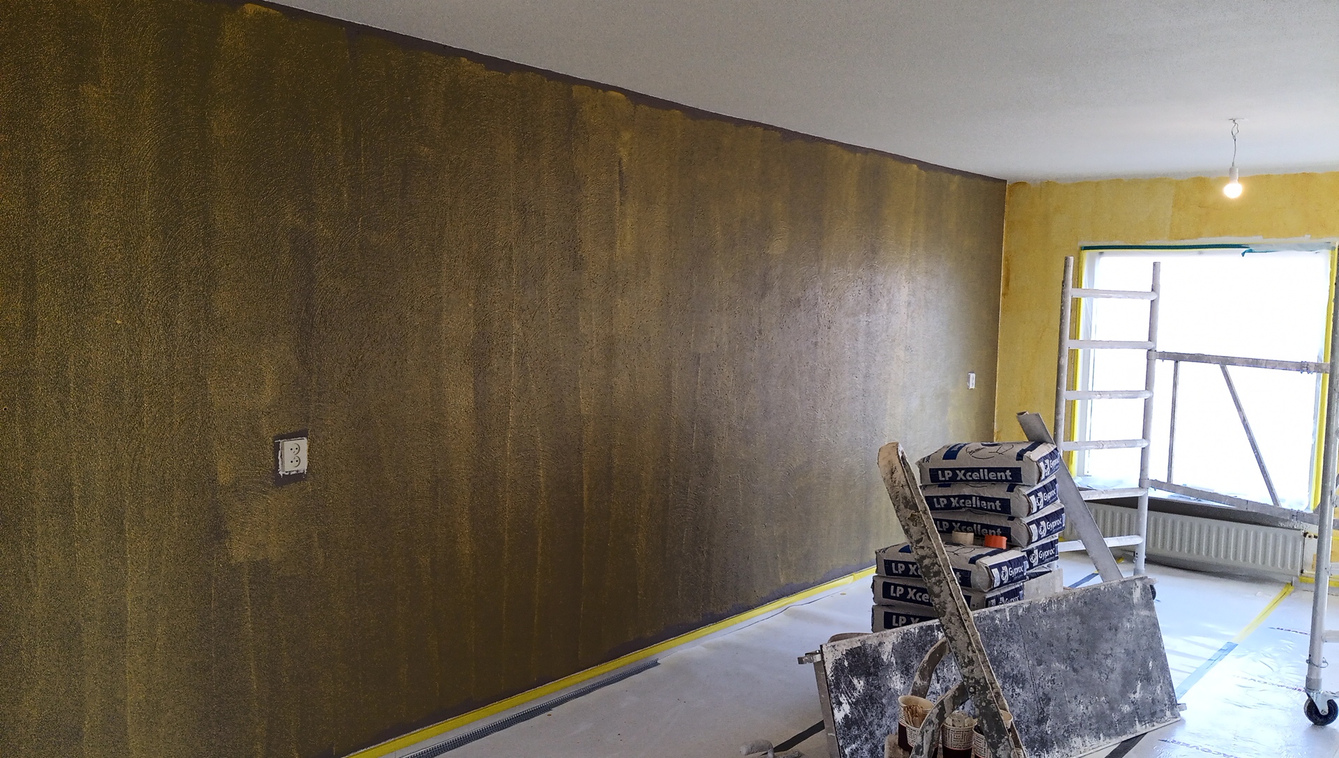 Lange wand tijdens het stucwerk, met zakken stuc op een steekwagen en een steiger bij het raam